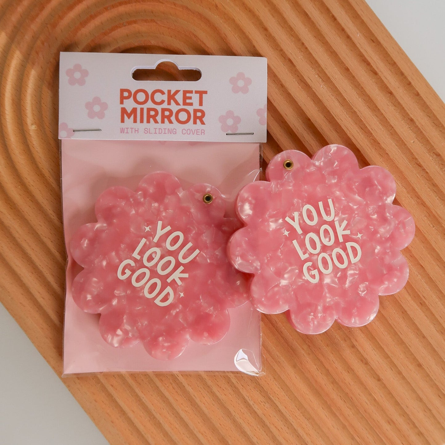 Pink flower-shaped pocket mirror with 'YOU LOOK GOOD' text on a wooden surface