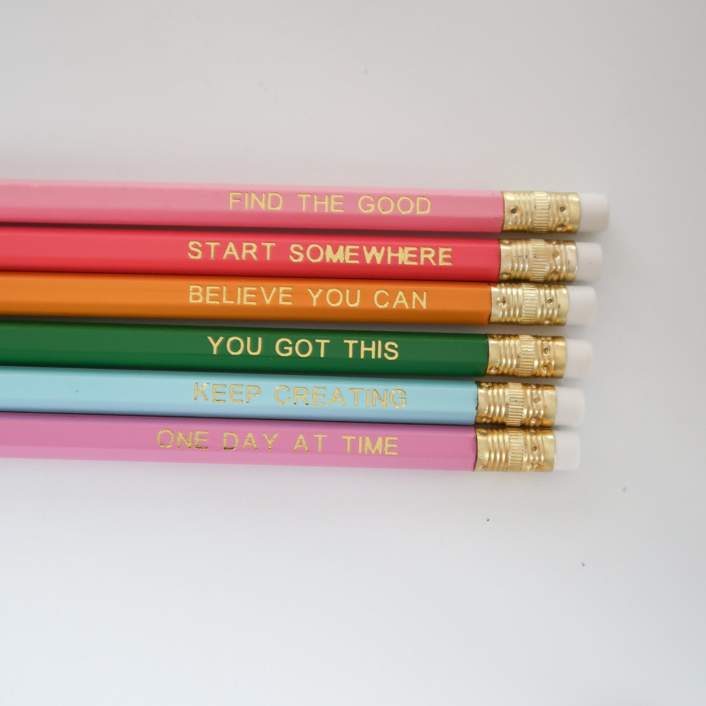Six colorful pencils arranged horizontally on a white background, each printed with a positive phrase in gold foil and finished with white erasers and gold ferrules.