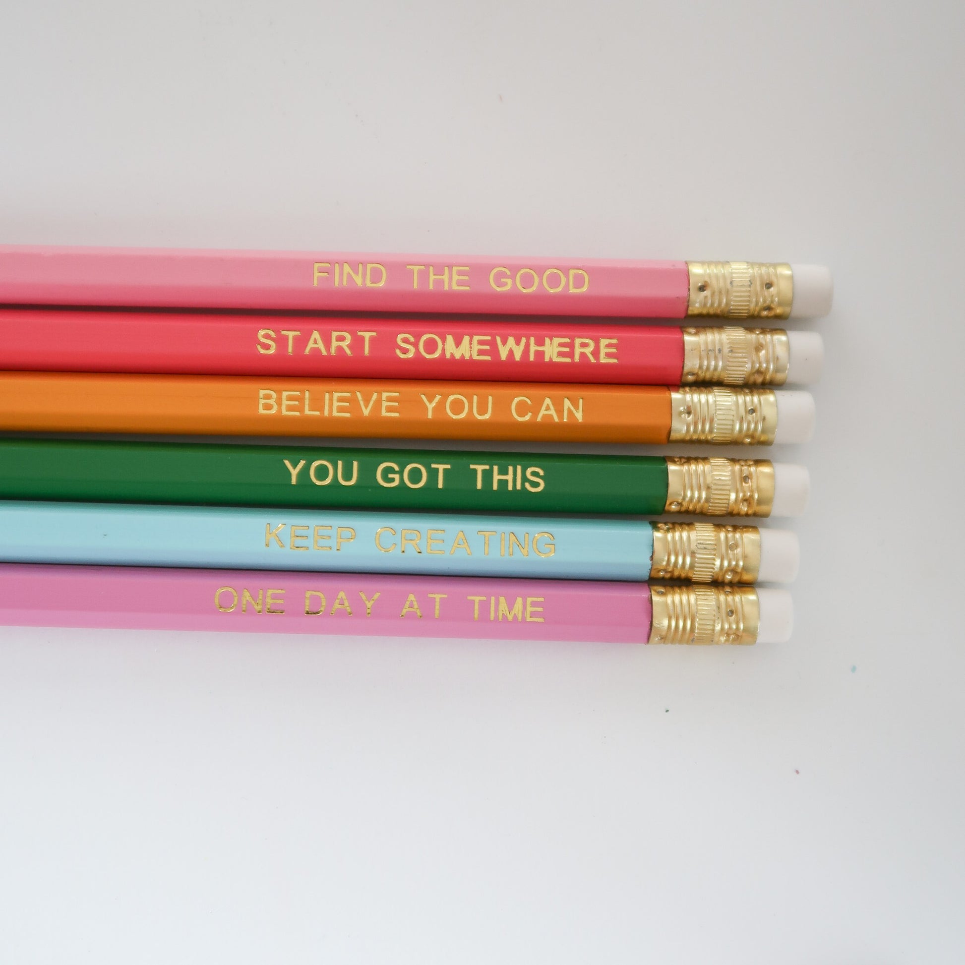 Six colorful pencils arranged horizontally on a white background, each printed with a positive phrase in gold foil and finished with white erasers and gold ferrules.