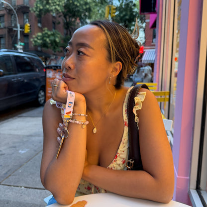A woman with her hair in a bun sits at a white table outside a shop, wearing a floral strapless top and multiple colorful bracelets. The background shows a city street with buildings, trees, and a parked car.