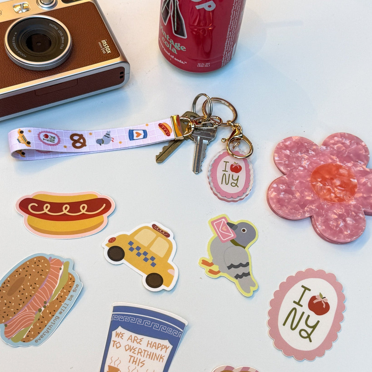 A collection of colorful die-cut stickers and a matching keychain strap laid out on a white surface, alongside a retro camera, a red soda can, and a pink flower-shaped object. The stickers feature New York-themed designs including a hot dog, taxi, pigeon, croissant, coffee cup, and 'I <3 NY' motifs.