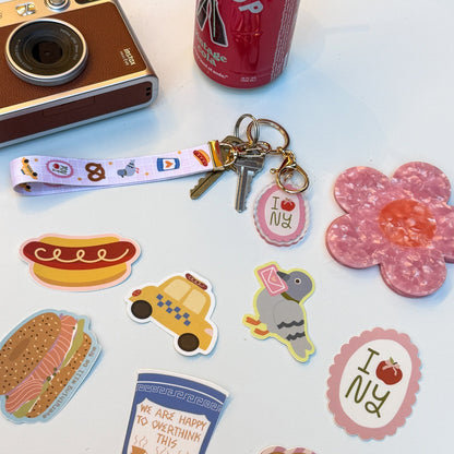 A collection of colorful die-cut stickers and a matching keychain strap laid out on a white surface, alongside a retro camera, a red soda can, and a pink flower-shaped object. The stickers feature New York-themed designs including a hot dog, taxi, pigeon, croissant, coffee cup, and 'I <3 NY' motifs.