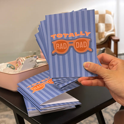 Hand holding a stack of blue cards with 'Totally Rad Dad' text in a casual indoor setting.