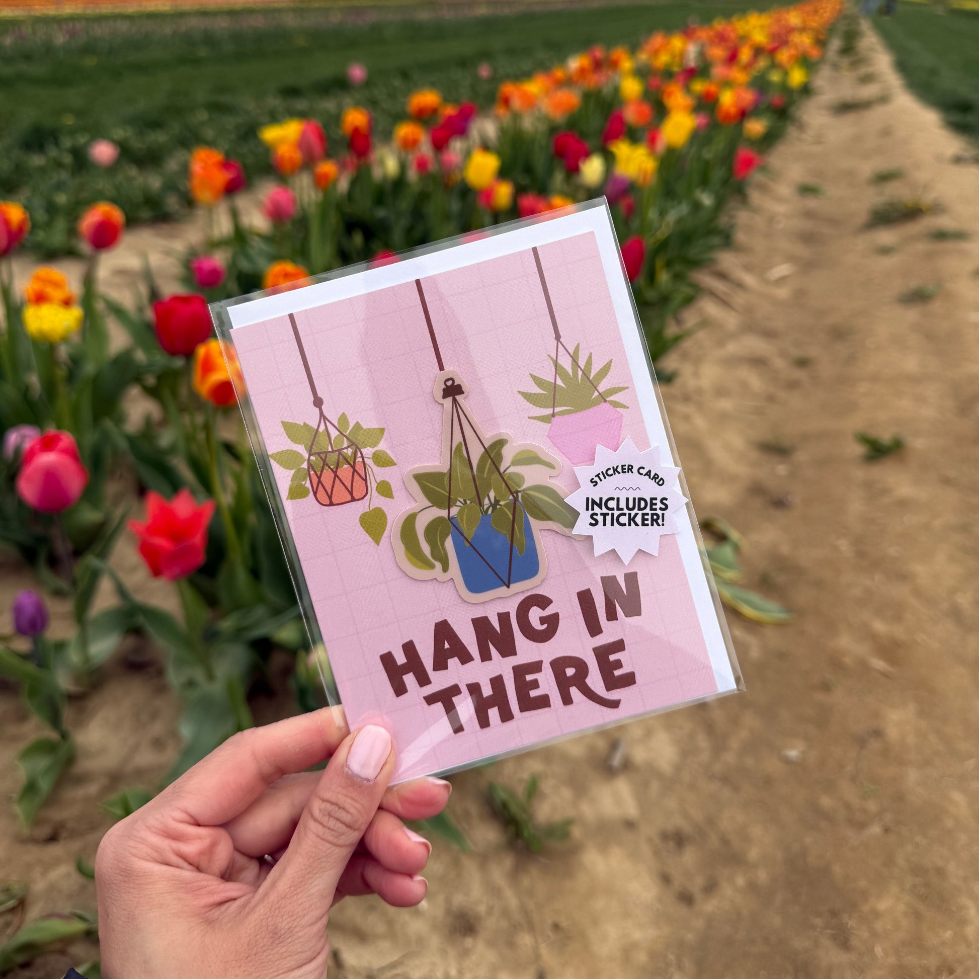 Hand holding a card with 'Hang in there' message in front of a colorful flower field.
