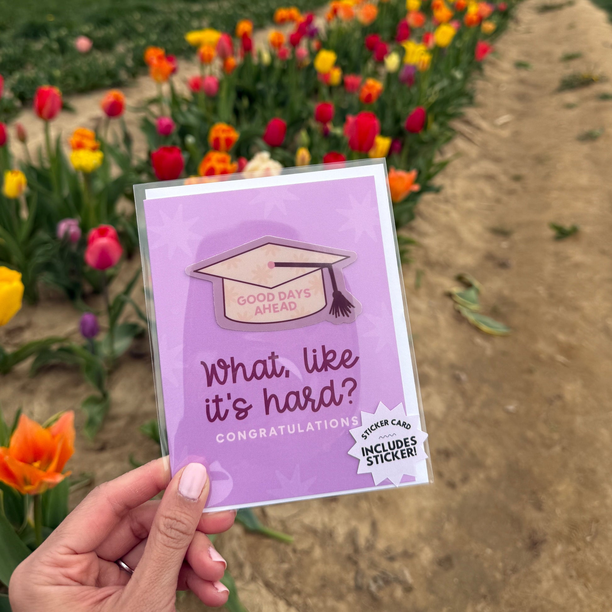 Hand holding a purple card with text and an illustration of a graduation cap against a background of flowers.