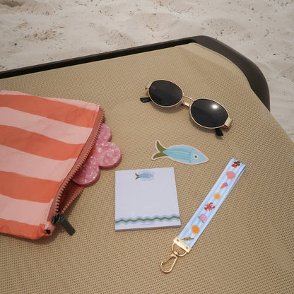 Sunglasses, notepad, fish-shaped magnet, and floral keychain on a textured surface